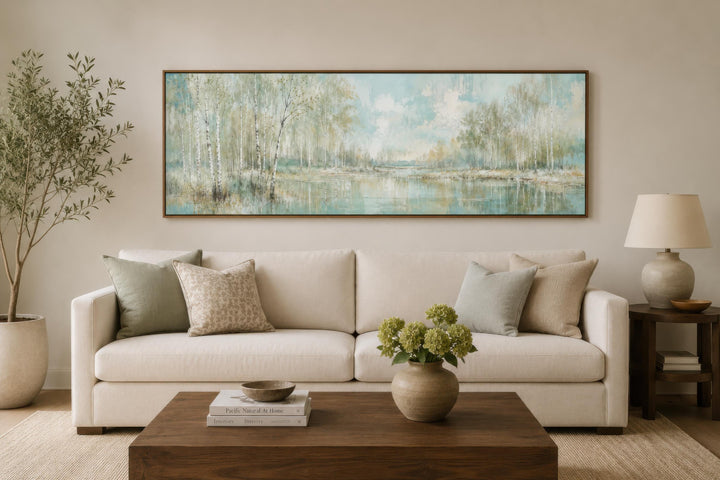 Long horizontal canvas print of a misty birch tree forest over a sofa in a neutral living room.