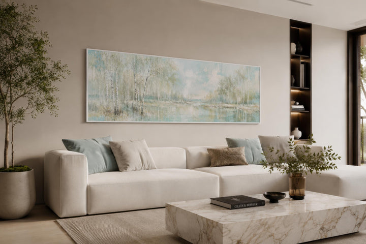 Long horizontal canvas print of a misty birch tree forest over a sofa in a neutral living room.