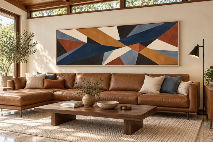 Long horizontal geometric abstract canvas wall art in earth tones above a brown leather sofa.