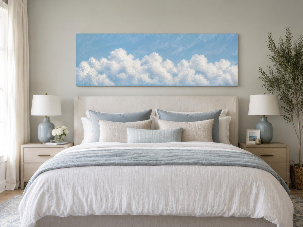 Long horizontal canvas painting of white clouds on a blue sky above a bed in a coastal bedroom.