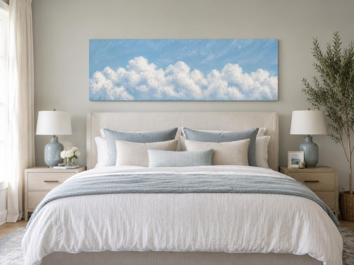 Long horizontal canvas painting of white clouds on a blue sky above a bed in a coastal bedroom.