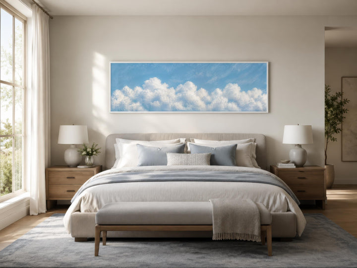 Long horizontal white cloud painting on blue sky canvas above a bed in a coastal bedroom.
