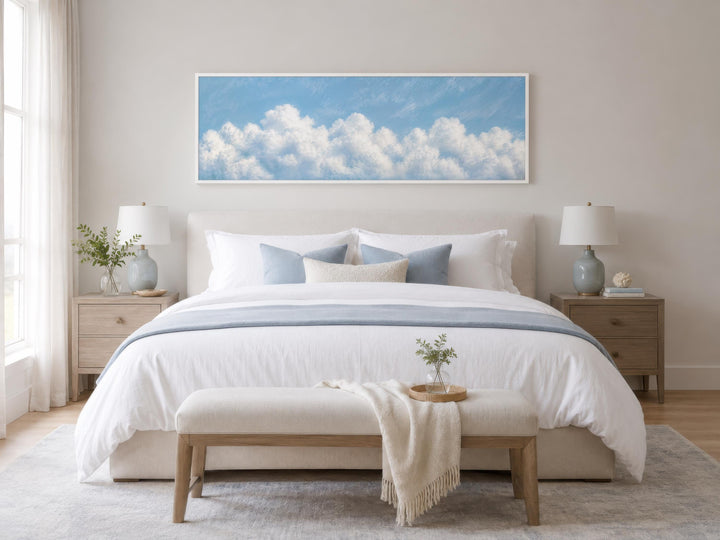 Long horizontal white cloud painting on blue sky canvas above a bed in a coastal bedroom.