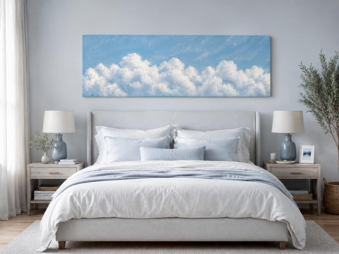 Long horizontal white cloud painting on blue sky canvas above a bed in a coastal bedroom.