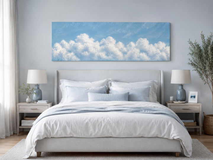 Long horizontal white cloud painting on blue sky canvas above a bed in a coastal bedroom.