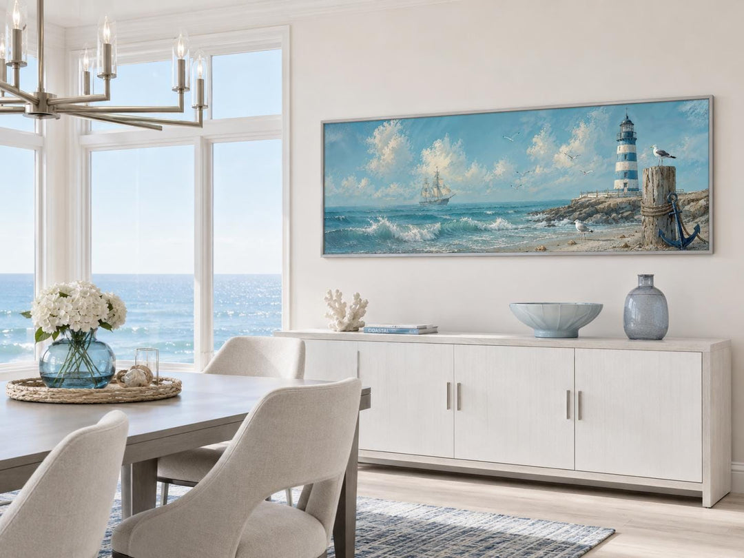 Long nautical canvas print of a sailing ship and lighthouse above a white sideboard in a beach room.