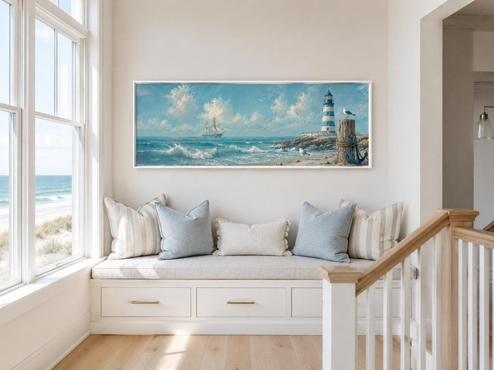 Long sailing ship and lighthouse canvas print in a coastal window nook with blue and white pillows.