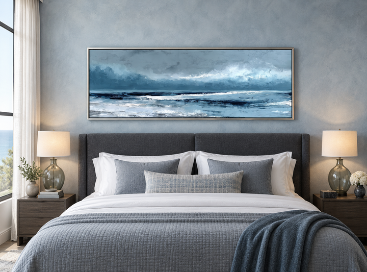 Long horizontal light blue ocean canvas wall art in a silver frame above a bed in a bedroom.