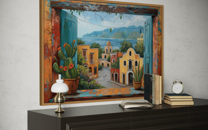 Mexican Village Window View Framed Canvas Wall Art on a dark wood dresser with books and a lamp.