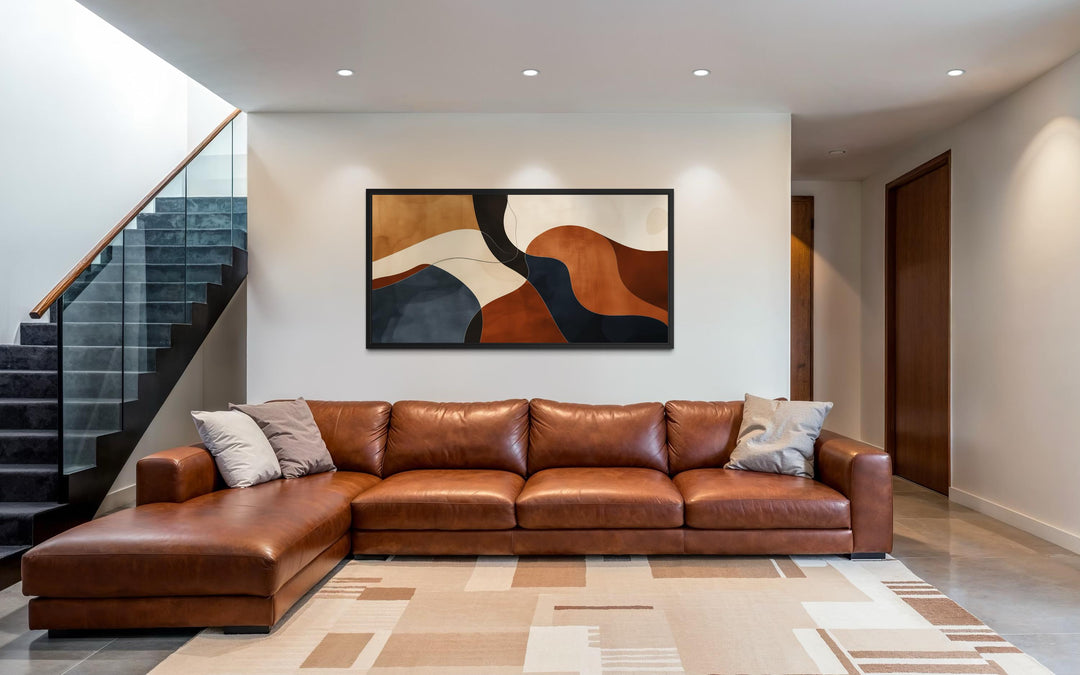 Mid Century abstract canvas print with curved shapes in earth tones above a brown leather sofa.
