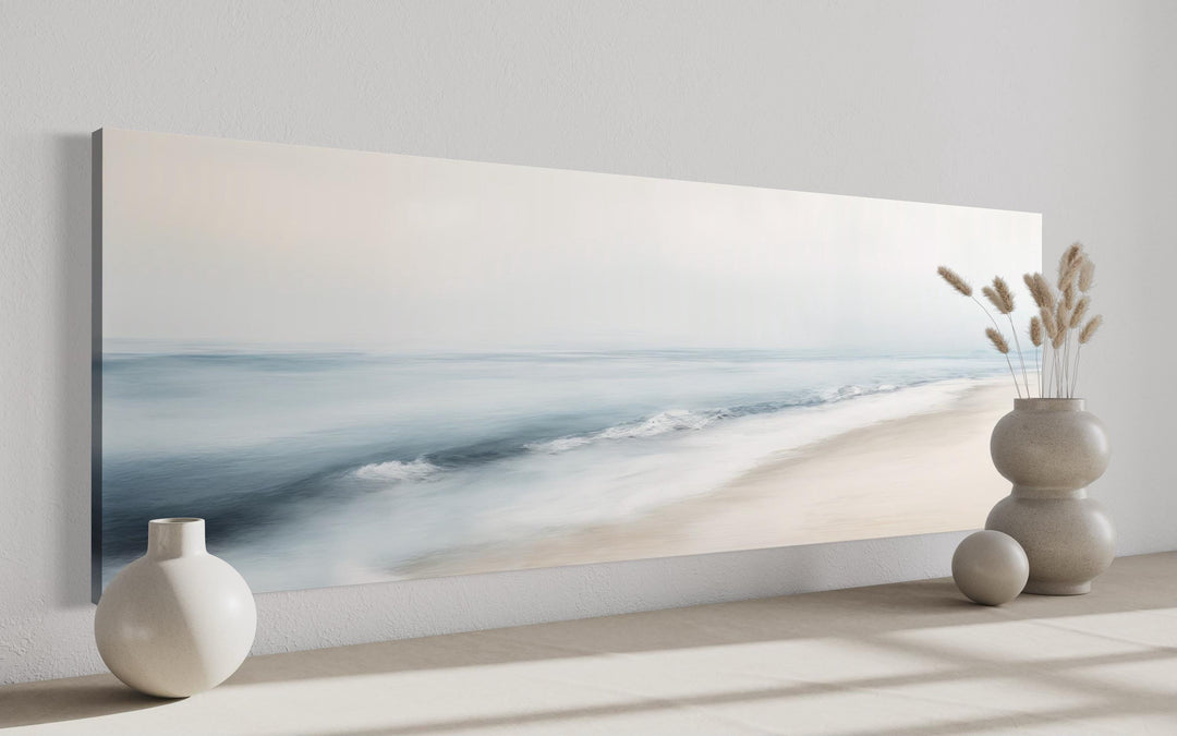 Panoramic minimalist watercolor ocean beach wall art in soft blue and gray tones on a white wall.