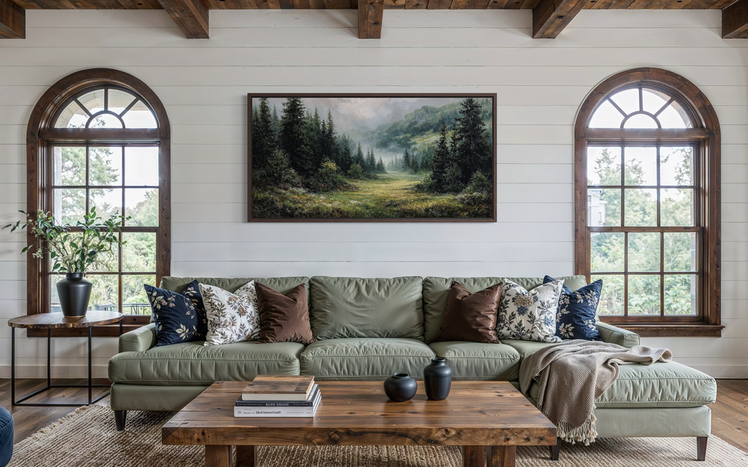 A panoramic canvas painting of a misty mountain meadow and pine forest hangs above a green sofa.