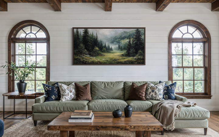 A panoramic canvas painting of a misty mountain meadow and pine forest hangs above a green sofa.