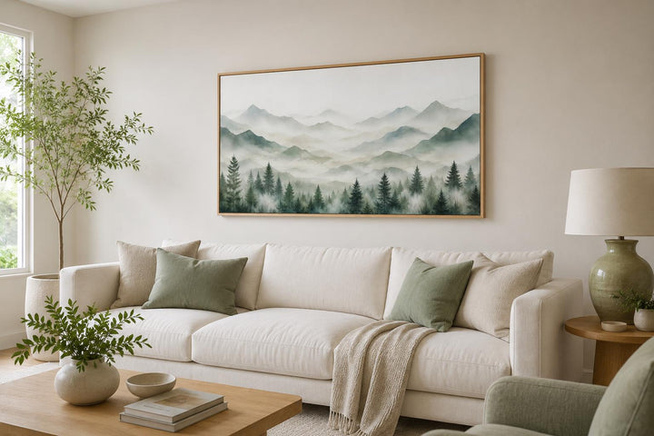 Misty mountain painting with sage green pine trees on canvas above a white sofa in a living room.