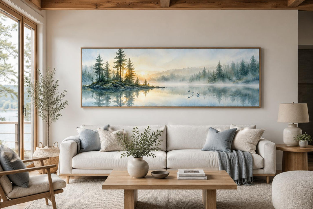 Panoramic misty pine forest and serene lake canvas print above a sofa in a neutral living room.