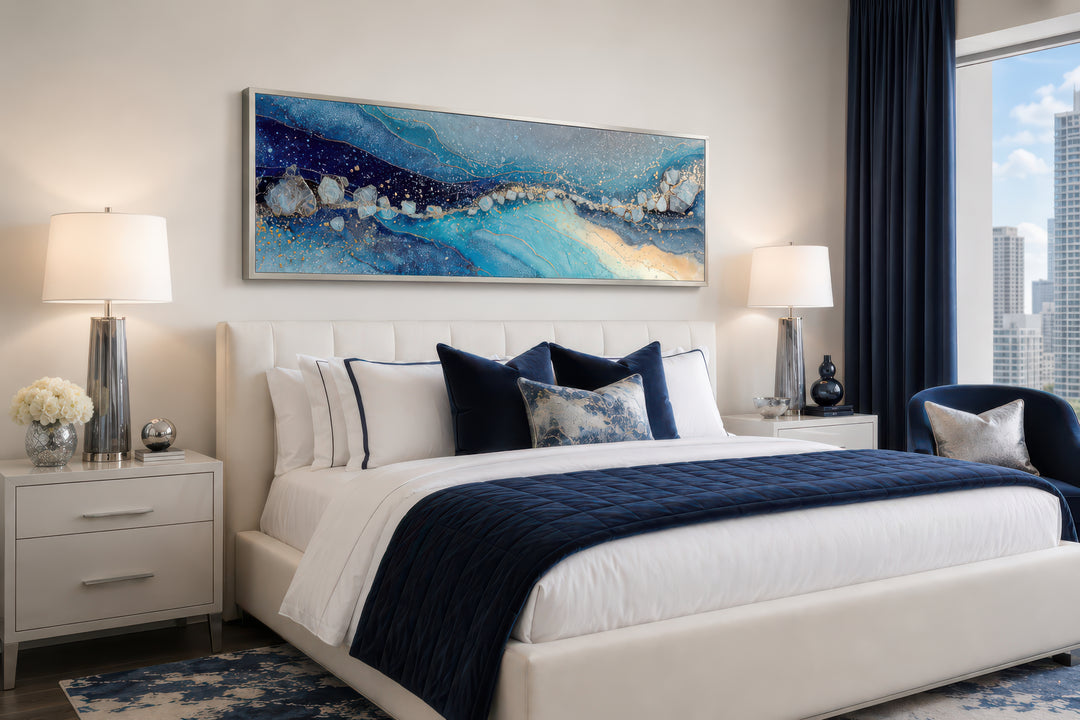 Modern long horizontal blue and navy abstract canvas art hanging above a white bed in a bedroom.