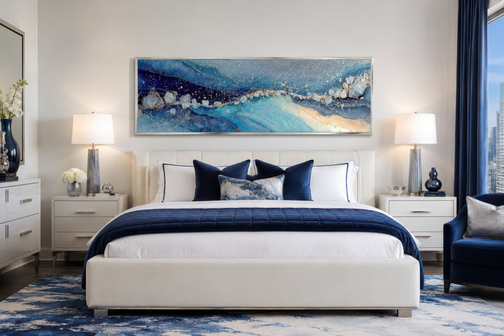 Modern long horizontal blue and navy abstract canvas art hanging above a white bed in a bedroom.