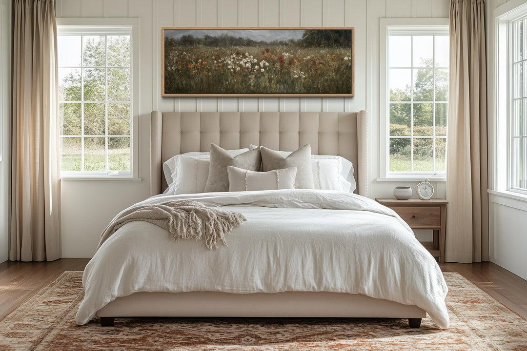 A long horizontal canvas of a muted wildflower field hangs above a bed in a neutral bedroom.