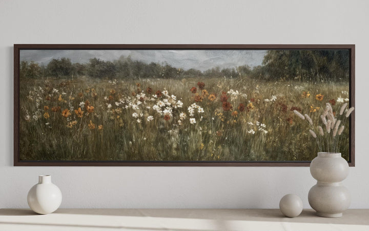 A long horizontal canvas painting of a muted wildflower field in a dark wood frame above a shelf.