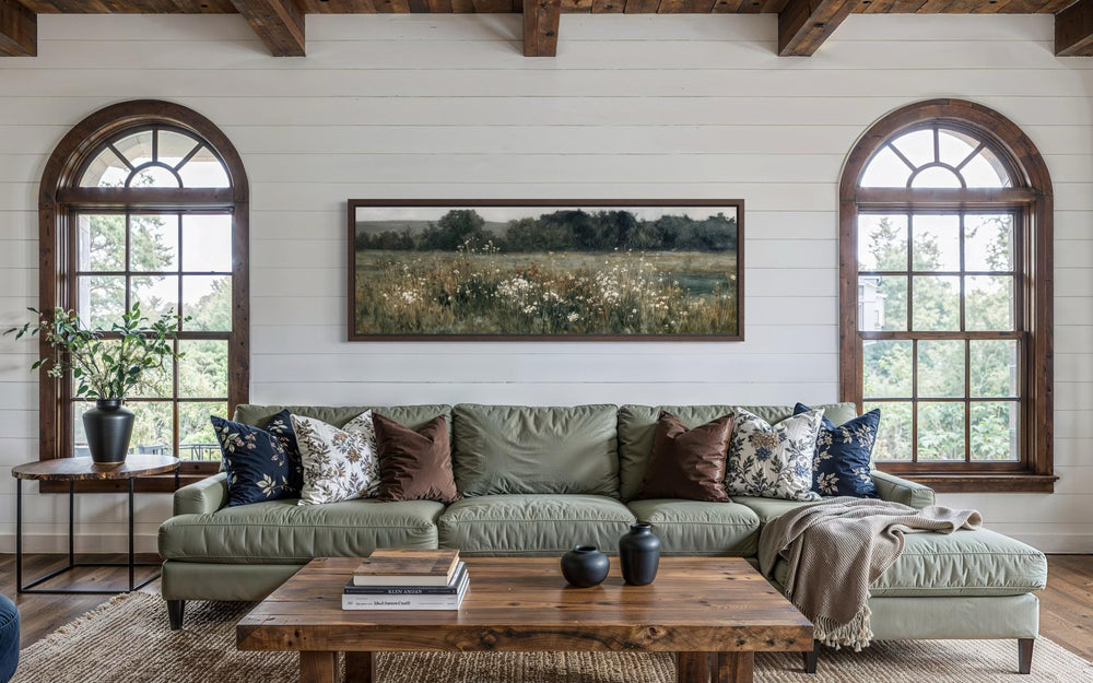 A long, narrow horizontal painting of a moody wildflower field hangs above a green sofa.