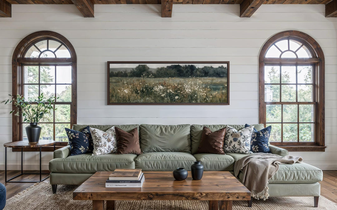 A long, narrow horizontal painting of a moody wildflower field hangs above a green sofa.