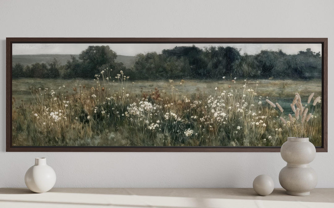A long horizontal painting of a moody wildflower field in a dark wood frame above a neutral shelf.