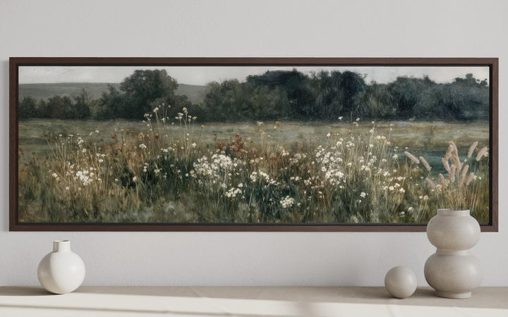 A long horizontal painting of a moody wildflower field in a dark wood frame above a neutral shelf.