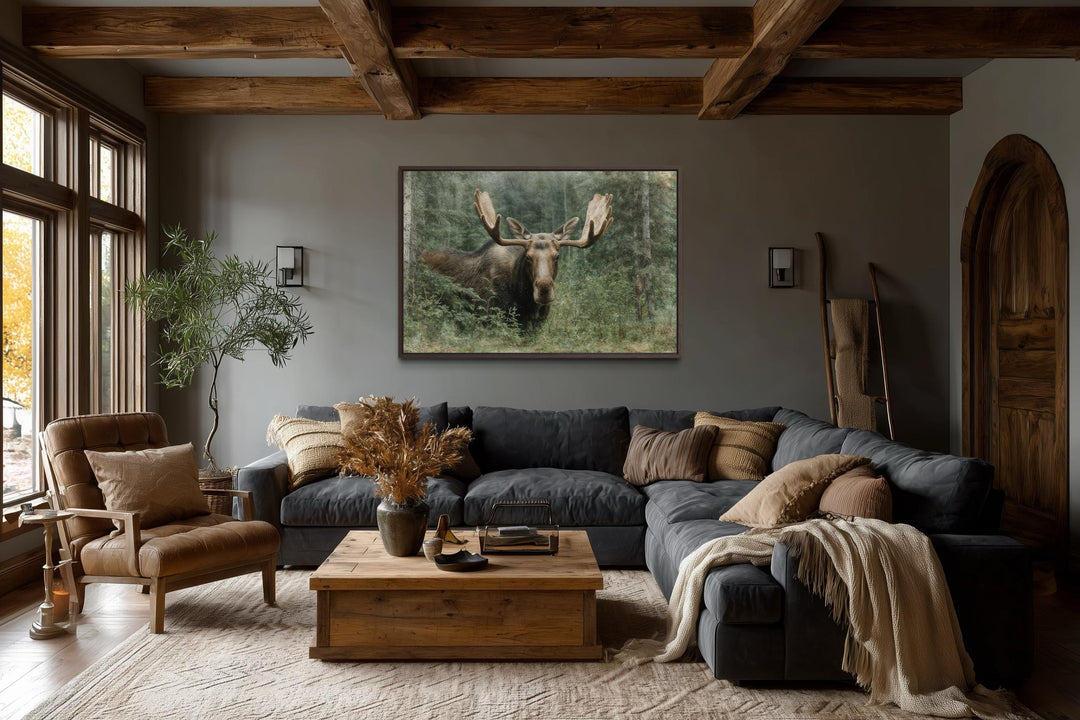 A rustic canvas painting of a moose in a forest hangs above a sofa in a cozy, wood-beamed lodge.