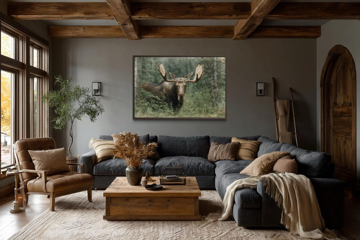 A rustic canvas painting of a moose in a forest hangs above a sofa in a cozy, wood-beamed lodge.