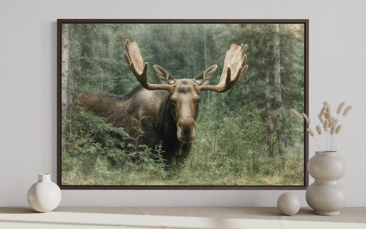 Moose in the forest wall art in a dark wood frame displayed above a shelf with decorative vases.