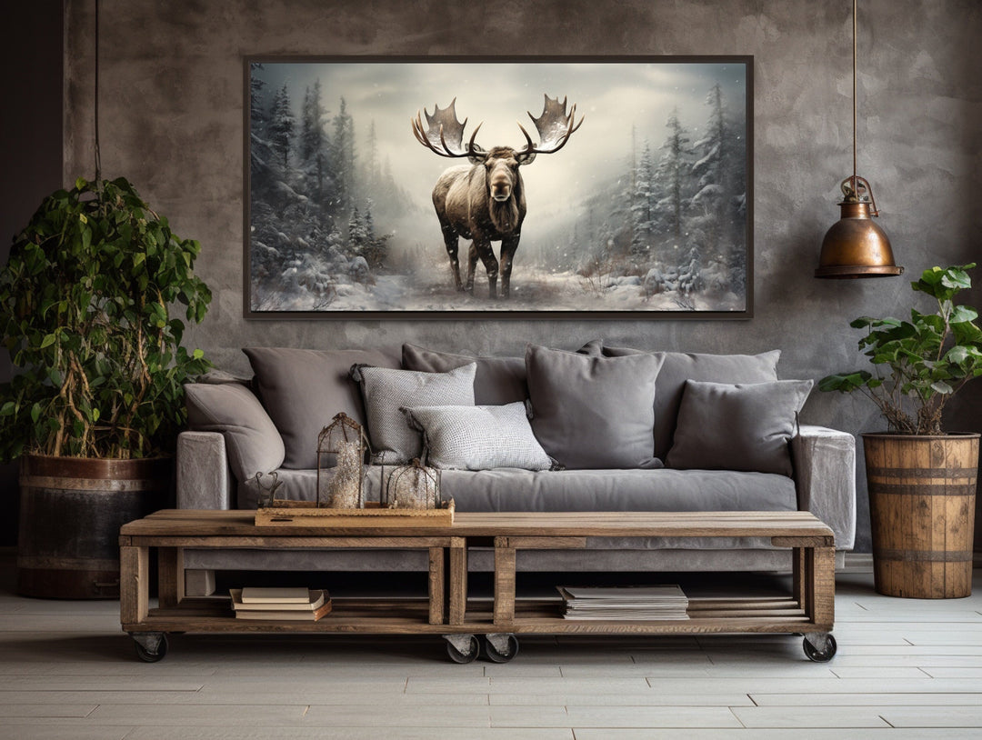 Framed canvas print of a moose in a snowy winter forest hanging above a gray sofa in a rustic room.