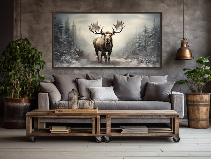 Framed canvas print of a moose in a snowy winter forest hanging above a gray sofa in a rustic room.