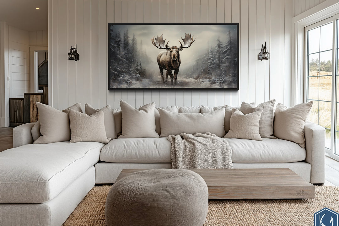 Framed canvas print of a moose in a snowy winter forest hanging above a white sectional sofa.