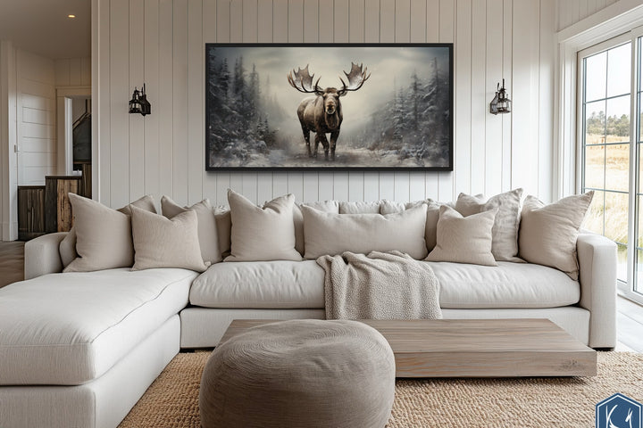 Framed canvas print of a moose in a snowy winter forest hanging above a white sectional sofa.