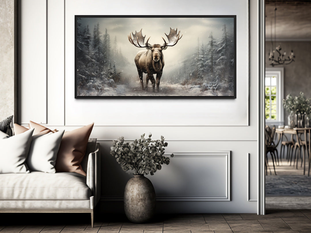 Framed canvas print of a moose in a snowy winter forest hanging above a sofa in a modern room.