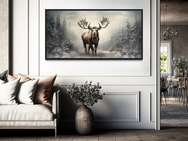 Framed canvas print of a moose in a snowy winter forest hanging above a sofa in a modern room.