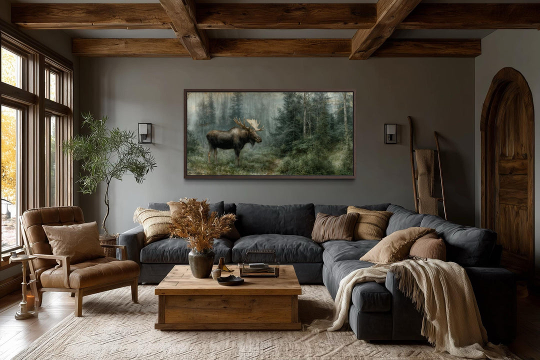 A large moose canvas print hangs in a rustic living room with dark wood beams and a leather chair.