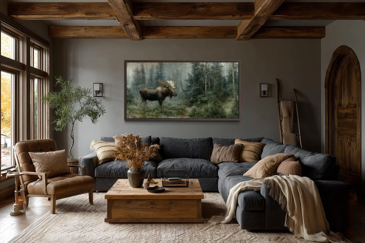 A large moose canvas print hangs in a rustic living room with dark wood beams and a leather chair.