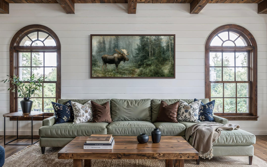 A large moose canvas print hangs on a white shiplap wall above a green sofa in a rustic living room.