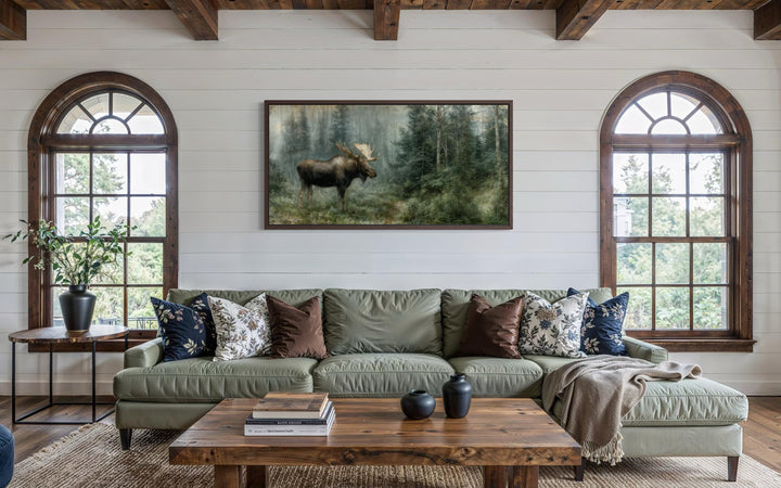 A large moose canvas print hangs on a white shiplap wall above a green sofa in a rustic living room.