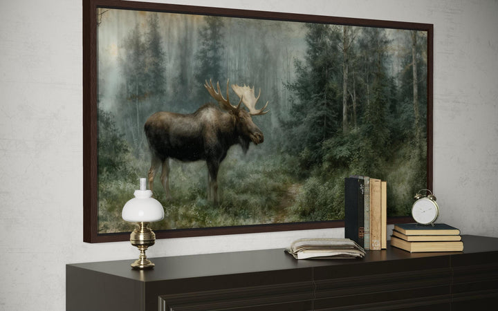 A rustic canvas print of a moose in a misty forest, framed and displayed above a dark wood dresser.