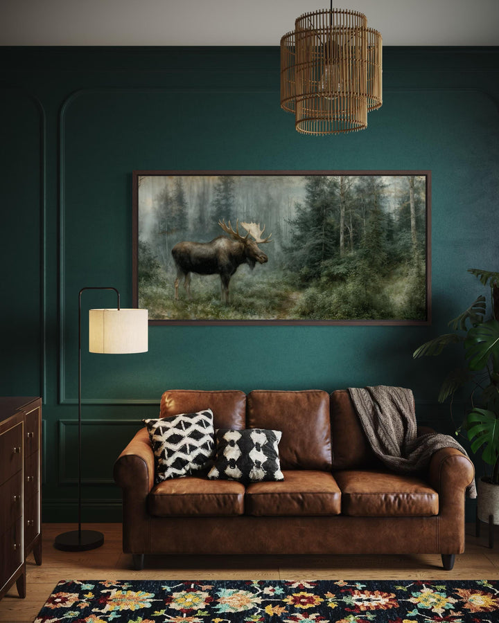 A large moose canvas print hangs on a dark green wall above a brown leather sofa in a rustic room.
