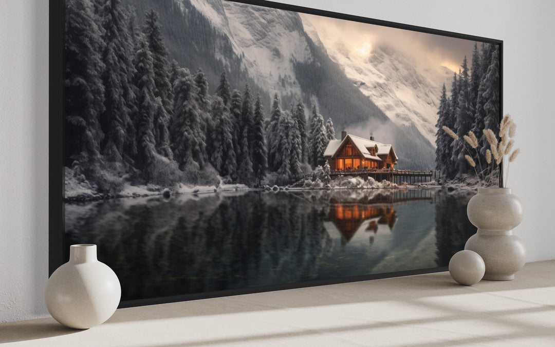 Panoramic framed canvas of a cozy winter cabin reflected in a lake with snowy mountains.