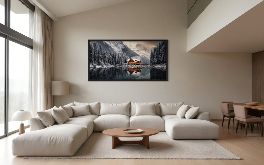 Framed panoramic canvas of a cozy winter cabin by a lake, hung above a modern sectional sofa.