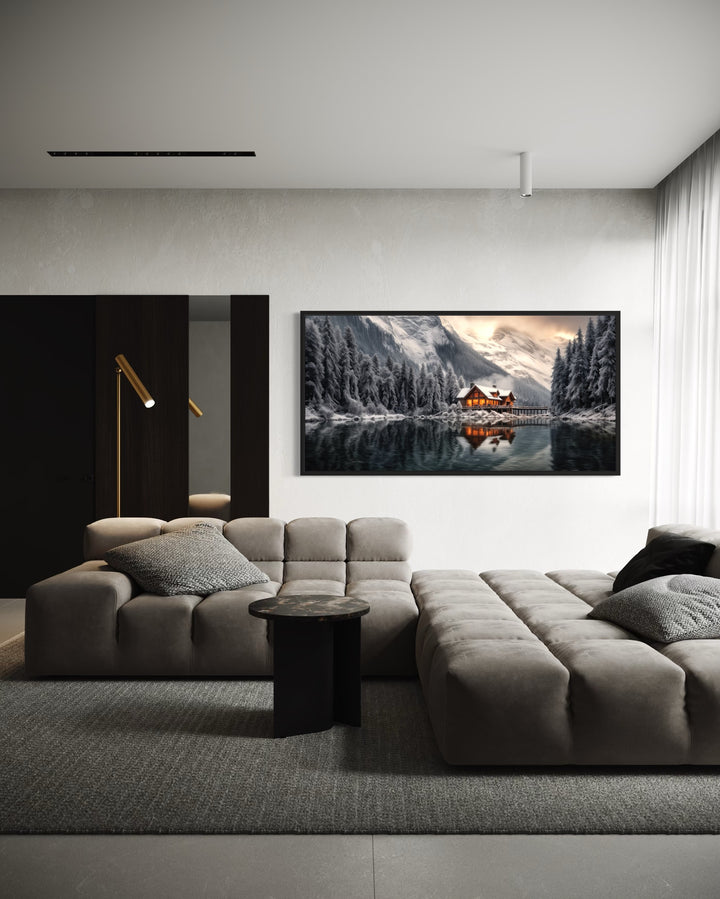 Framed panoramic canvas of a cozy winter cabin by a lake, hung above a modern grey sectional sofa.