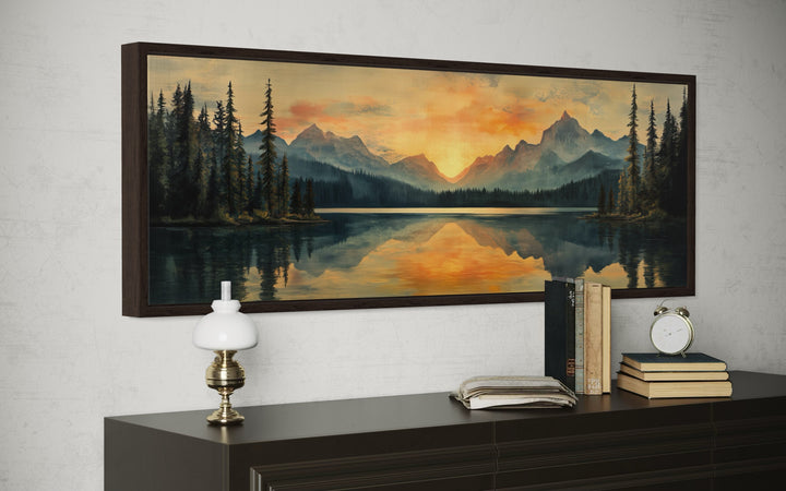 Panoramic mountain lake sunset wall art in a dark wood frame above a dresser with books and a lamp.