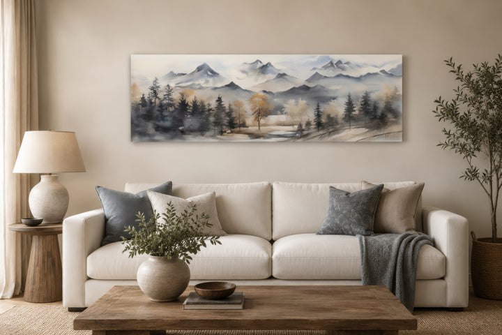 Horizontal framed canvas wall art of a watercolor mountain landscape in earth tones above a sofa.