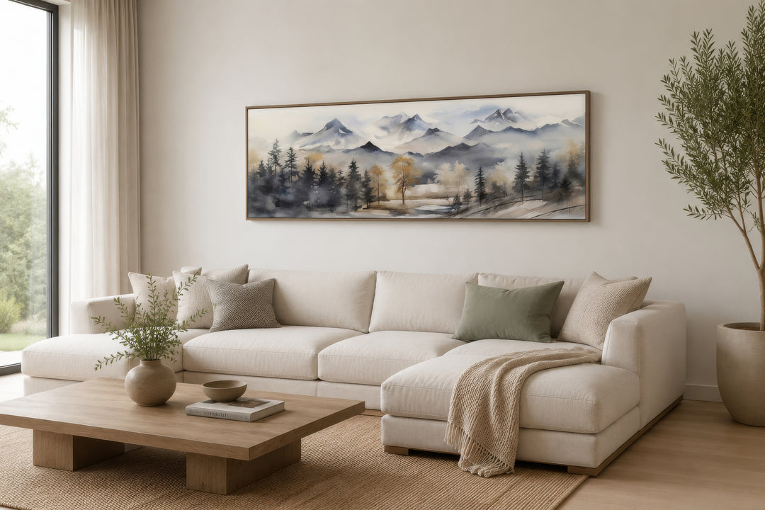 Horizontal framed canvas wall art of a watercolor mountain landscape in earth tones above a sofa.