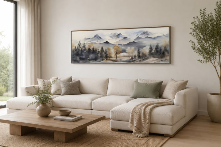 Horizontal framed canvas wall art of a watercolor mountain landscape in earth tones above a sofa.