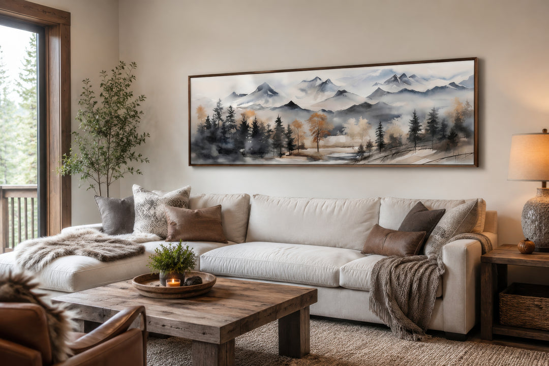 Horizontal framed canvas wall art of a watercolor mountain landscape in earth tones above a sofa.